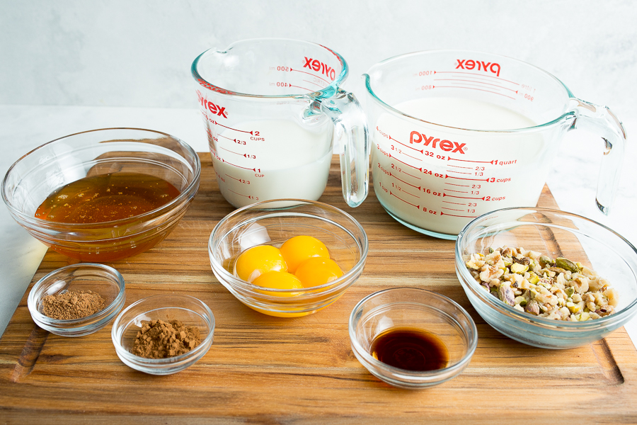 Ingredients for Baklava Ice Cream including milk, heavy cream, honey, cinnamon, nutmeg, egg yolks, vanilla extract, and nuts sit in individual bowls atop a wooden cutting board.