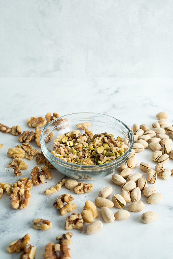 A clear bowl of leftover pistachios and walnuts on a white marble background.
